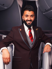 Virgin Atlantic cabin crew member standing in an aircraft aisle, wearing a burgundy uniform with a red tie and smiling warmly towards the camera. The crew member is leaning casually on the aircraft seats, with the cabin interior softly lit in the background