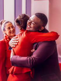 Virgin Atlantic crew members sharing a warm embrace inside an airport or cabin environment, with one smiling colleague visible in the background. The team are wearing Virgin Atlantic’s signature red and burgundy uniforms, capturing a welcoming and friendly customer service atmosphere.
